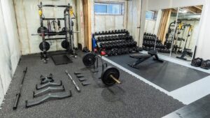 Ett välutrustat hemgym med hantlar, skivstänger och träningsbänk på svarta mattor.