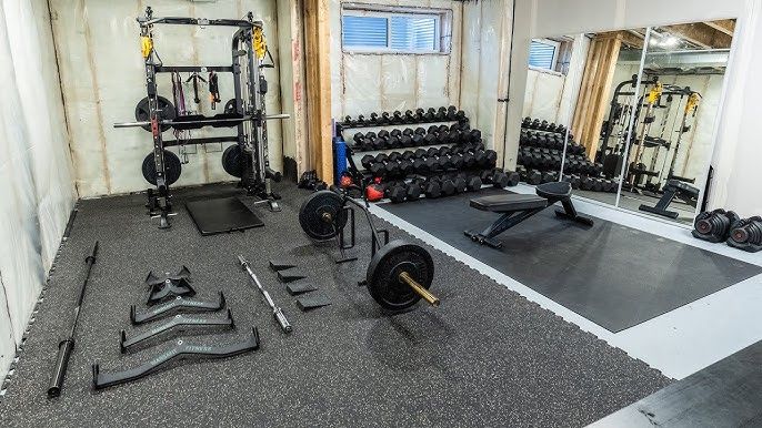Ett välutrustat hemgym med hantlar, skivstänger och träningsbänk på svarta mattor.