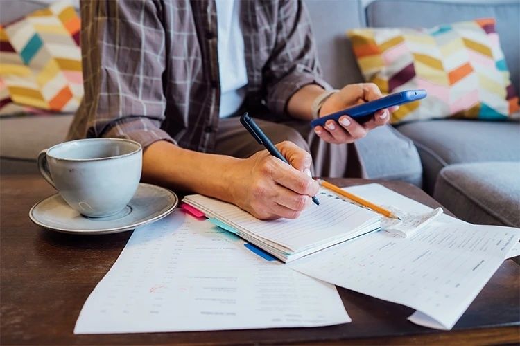 Person skriver i en anteckningsbok bredvid ekonomipapper och en kopp kaffe